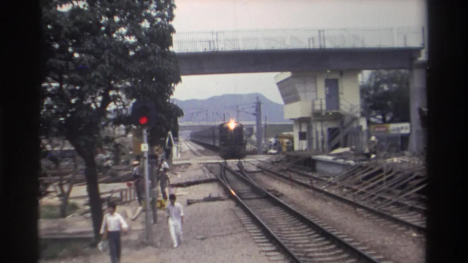 Final 8mm frame, Taiwan 1982: train at near distance with a very bright headlight; people walking alongside active tracks beneath concrete overpass, hills visible in background.