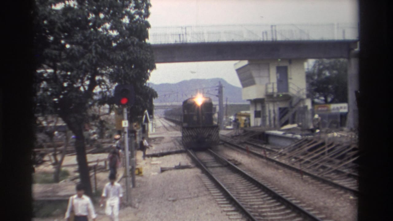 8mm home movie, Taiwan c.1982 -- locomotive with lit headlight approaching crossing, pedestrians in casual dress walking alongside tracks, concrete overpass visible above.