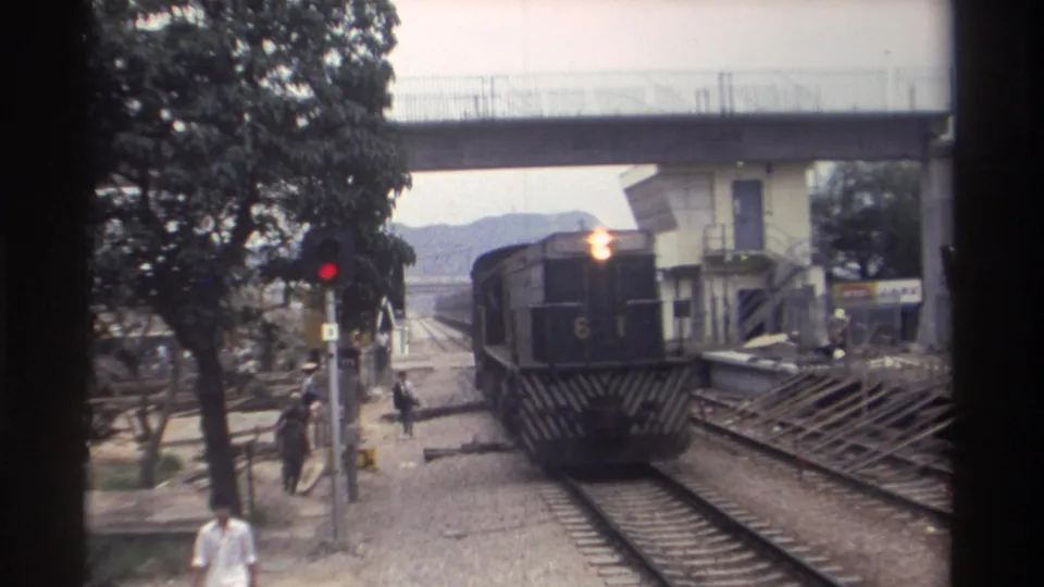 8mm home movie, Taiwan c.1982 -- diesel train moving under concrete footbridge at level crossing, red signal pole visible left, pedestrians and trees alongside.