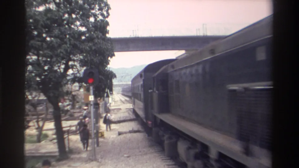8mm home movie, Taiwan c.1982 -- train carriages passing close to camera on right, pedestrians walking on trackside path left, mountains and concrete overpass in background.