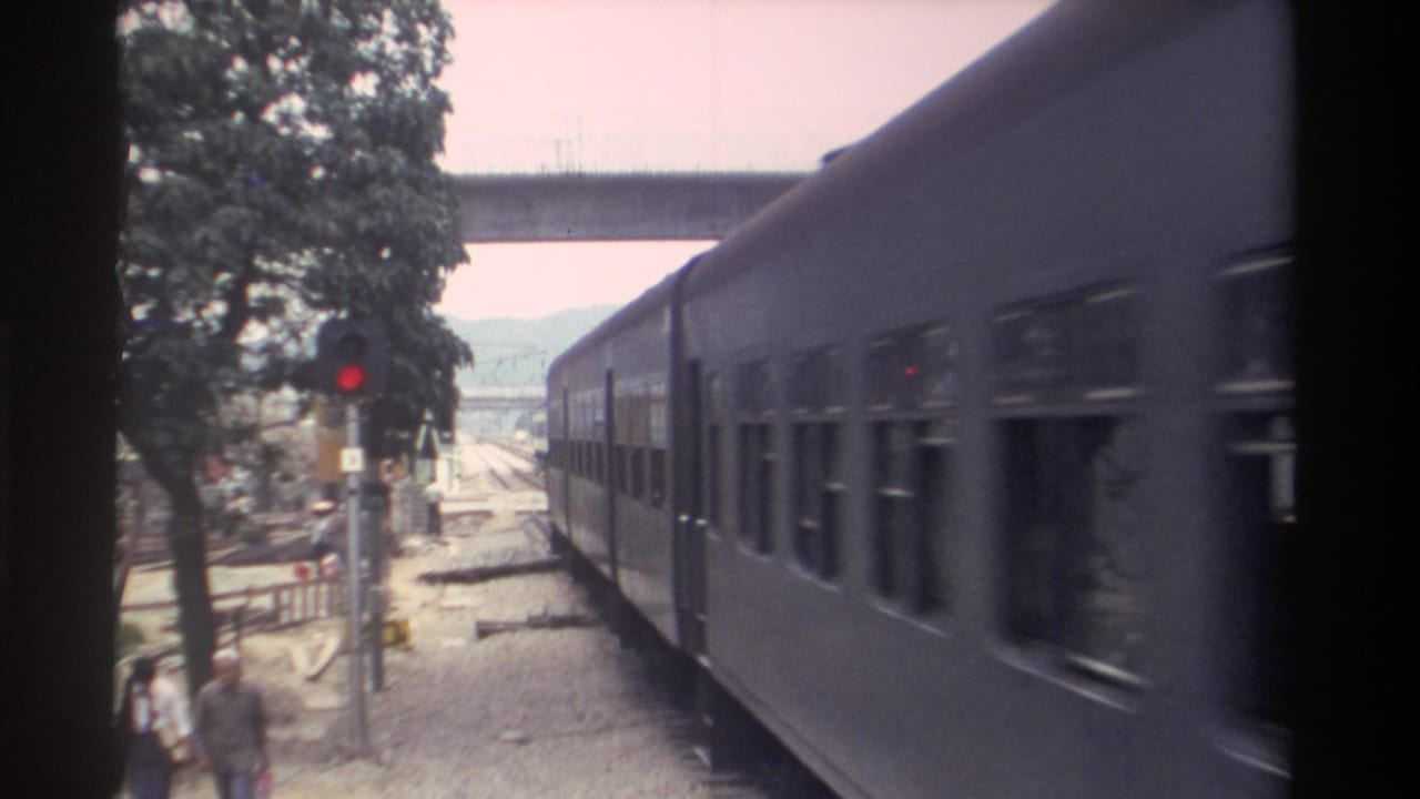8mm home movie frame, Taiwan 1982: a blue-grey passenger train passes a level crossing; red signal pole, concrete overpass, and pedestrian figure visible alongside the tracks.