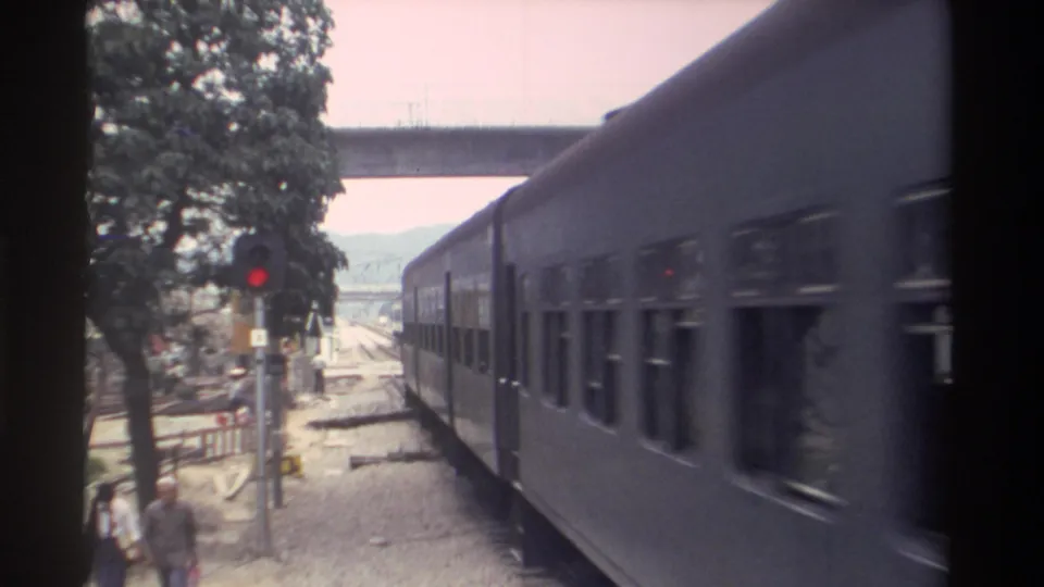 8mm home movie frame, Taiwan 1982: a blue-grey passenger train passes a level crossing; red signal pole, concrete overpass, and pedestrian figure visible alongside the tracks.