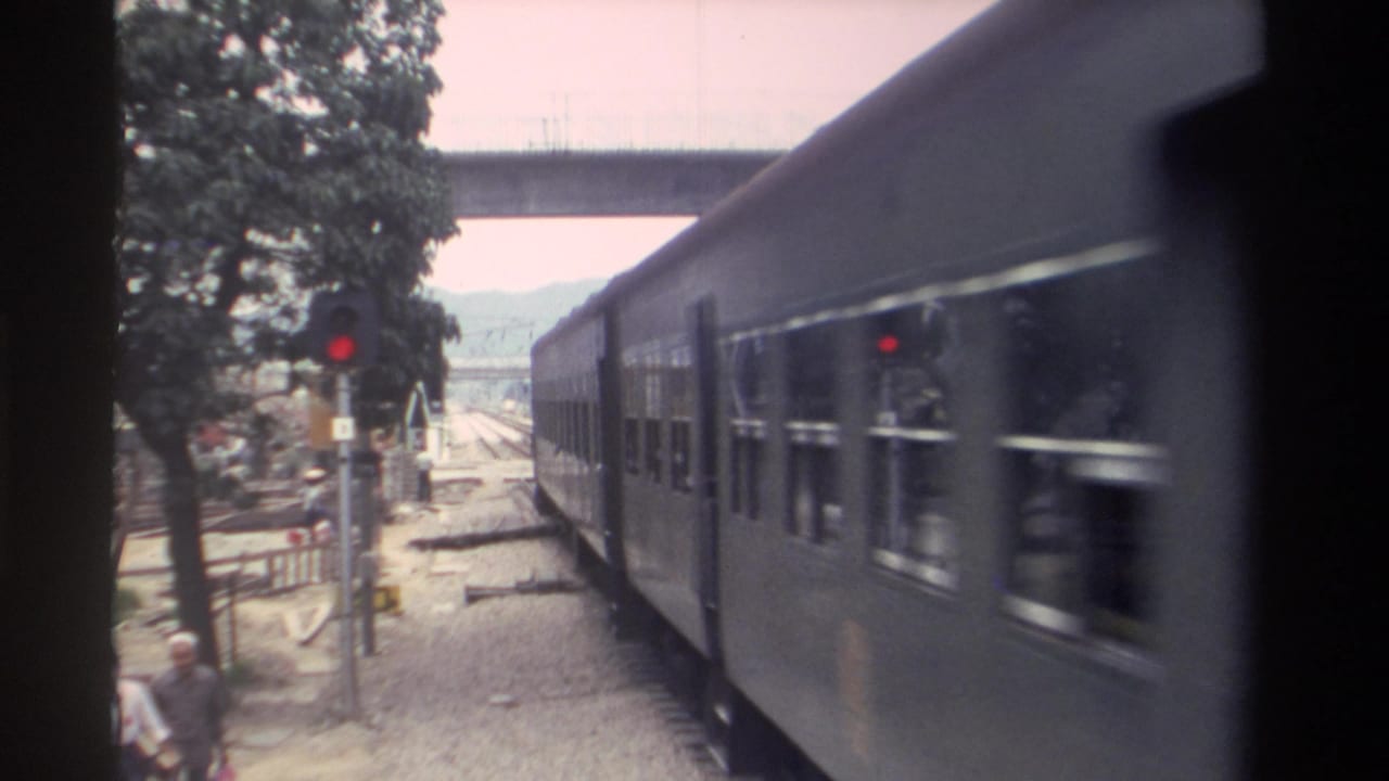 8mm footage, Taiwan 1982: passenger carriages fill the foreground as an adult pedestrian stands near the gravel trackside; red signal and concrete overpass frame the background.