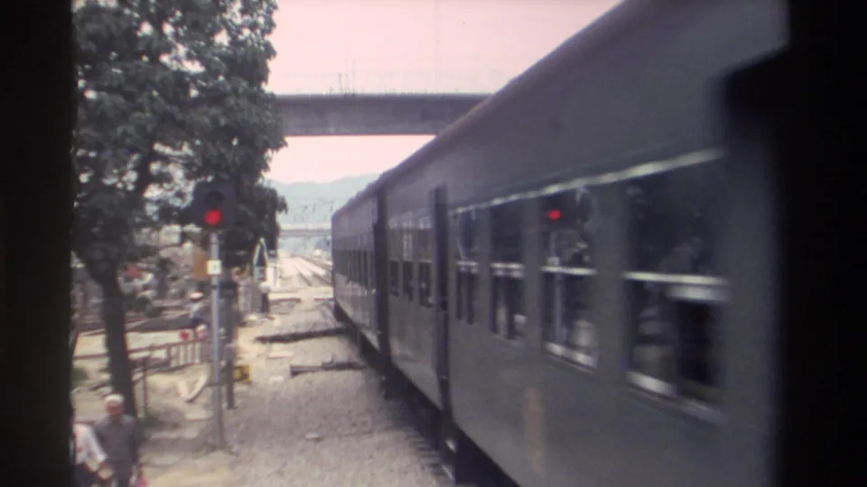 8mm footage, Taiwan 1982: passenger carriages fill the foreground as an adult pedestrian stands near the gravel trackside; red signal and concrete overpass frame the background.