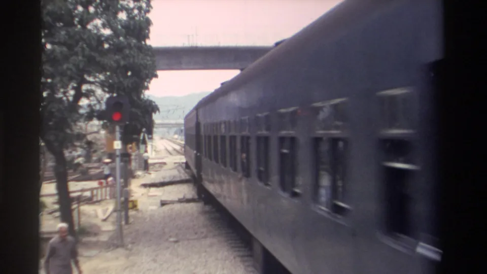 8mm home movie frame, Taiwan 1982: the blue-grey passenger train continues through the level crossing area as a pedestrian walks along the gravel beside the tracks.