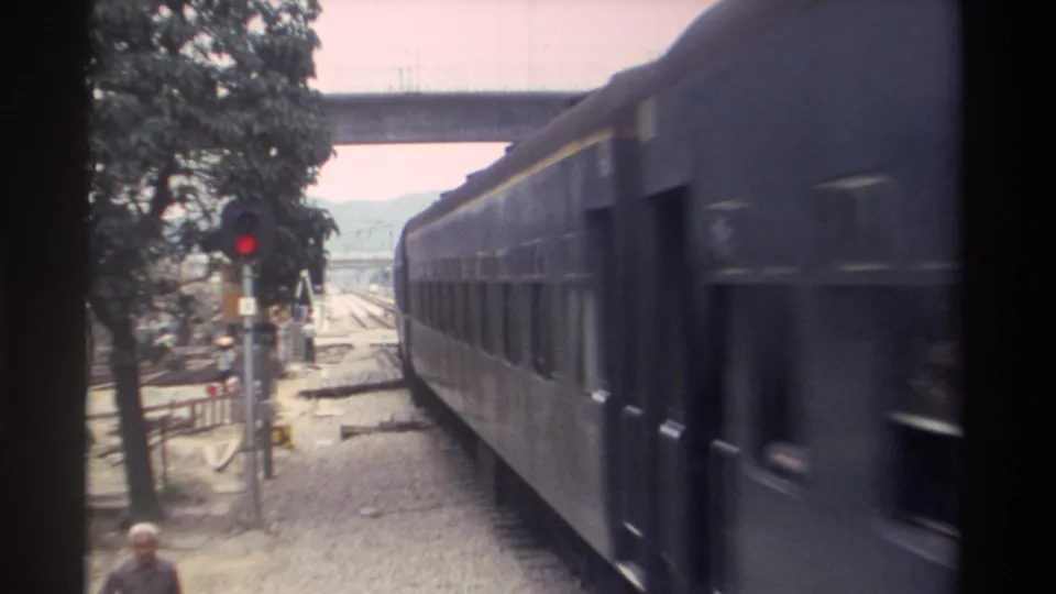 8mm footage, Taiwan 1982: passenger carriages occupy the right of frame as the train clears the level crossing; red signal pole, overpass, and a pedestrian figure remain visible.
