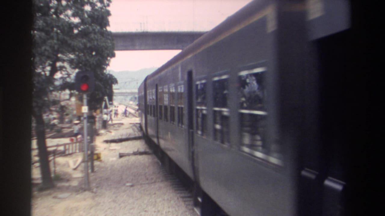 Passenger train continuing past a red signal light; pedestrians visible in the background beneath trees, concrete overpass at frame top.