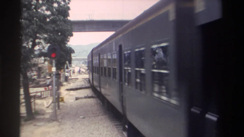 8mm view of a passenger train in motion near a railway signal in Taiwan, circa 1982; small groups of pedestrians visible in the background under mature trees.