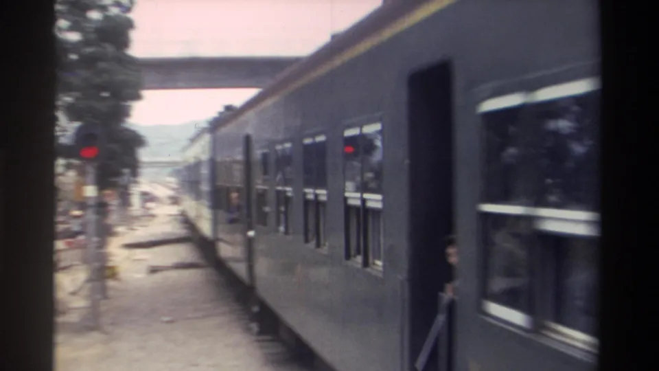Closing 8mm frame of a Taiwan passenger train passing from view, with red signal infrastructure, mature trees, and a solitary figure in the foreground, circa 1982.