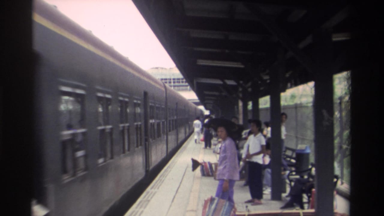 Covered railway platform in Taiwan circa 1982 with adult passengers, including a woman in lavender clothing, waiting alongside a dark blue passenger train.
