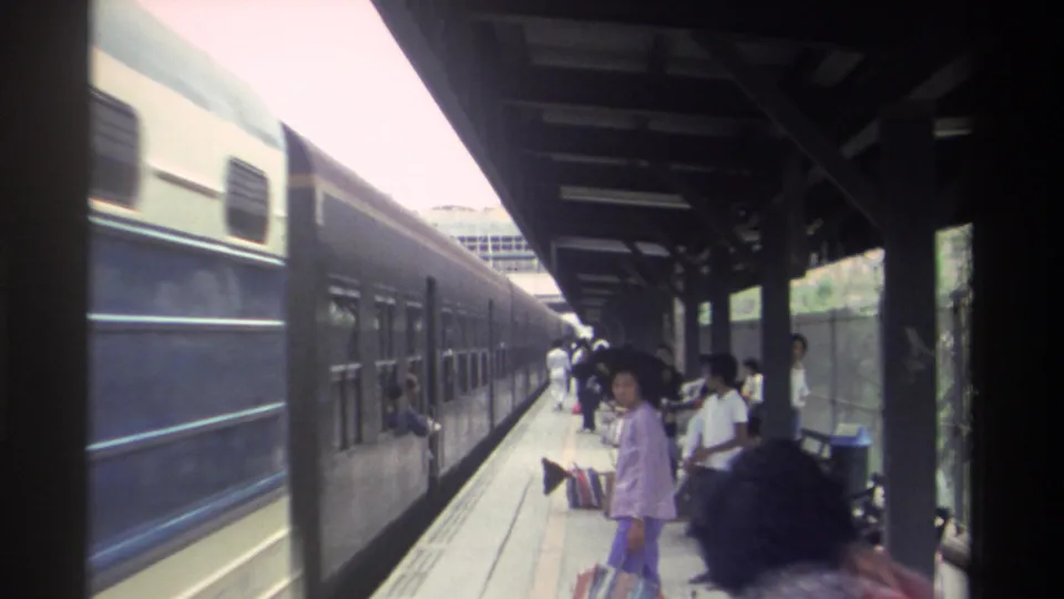 Taiwanese railway platform circa 1982: dark blue passenger train alongside a green-tiled canopy, casually dressed adult passengers including a woman in lavender flared trousers.