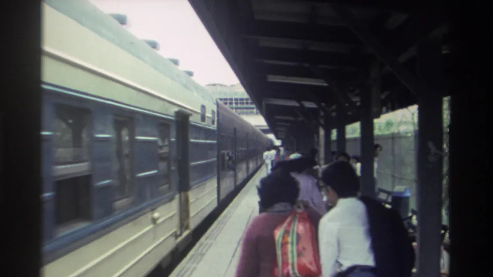 Taiwanese railway station platform circa 1982; passengers with a colorful striped bag stand in the foreground as a dark blue train stands alongside under a green-roofed canopy.