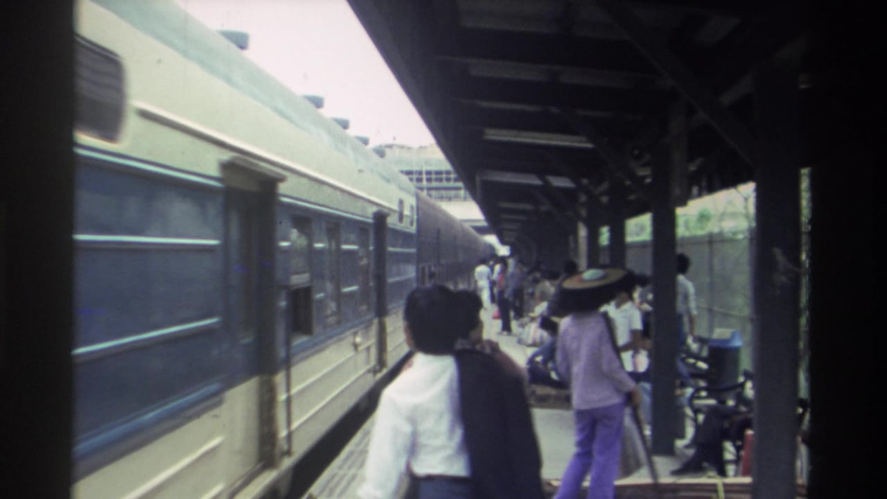 Passengers walking and standing on a covered timber-post platform alongside blue train carriages at a Taiwan railway station, circa 1982.