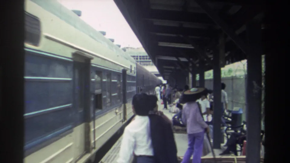 Passengers walking and standing on a covered timber-post platform alongside blue train carriages at a Taiwan railway station, circa 1982.