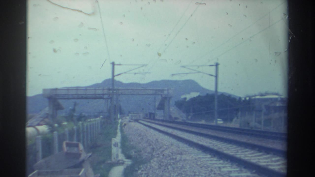 Train-forward POV with converging electrified tracks, a concrete overpass structure, and hazy mountains in the background, Taiwan, circa 1982.
