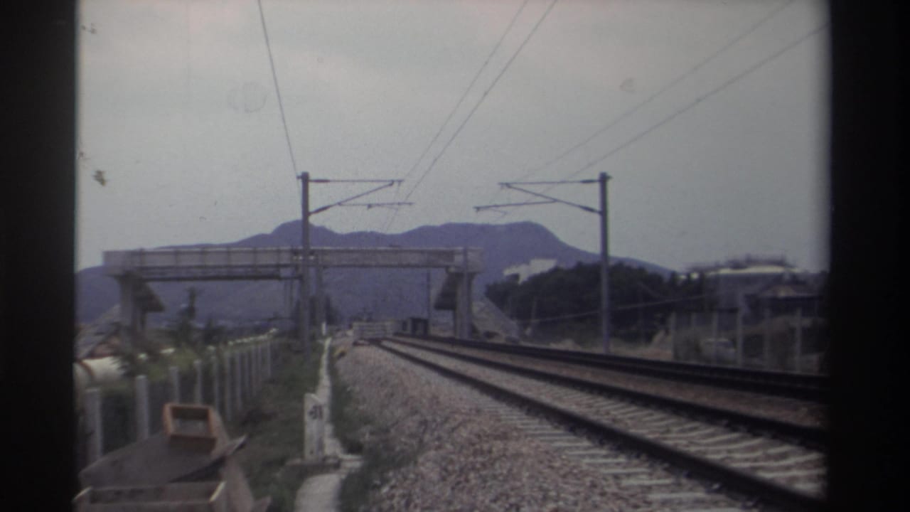 POV from moving train along electrified railway with concrete pedestrian overbridge and hazy mountains, Taiwan, 1982, Super 8 film.