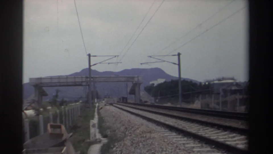 Continuation of train journey along electrified tracks; overbridge and mountain backdrop shift as train moves, Taiwan, 1982, Super 8.
