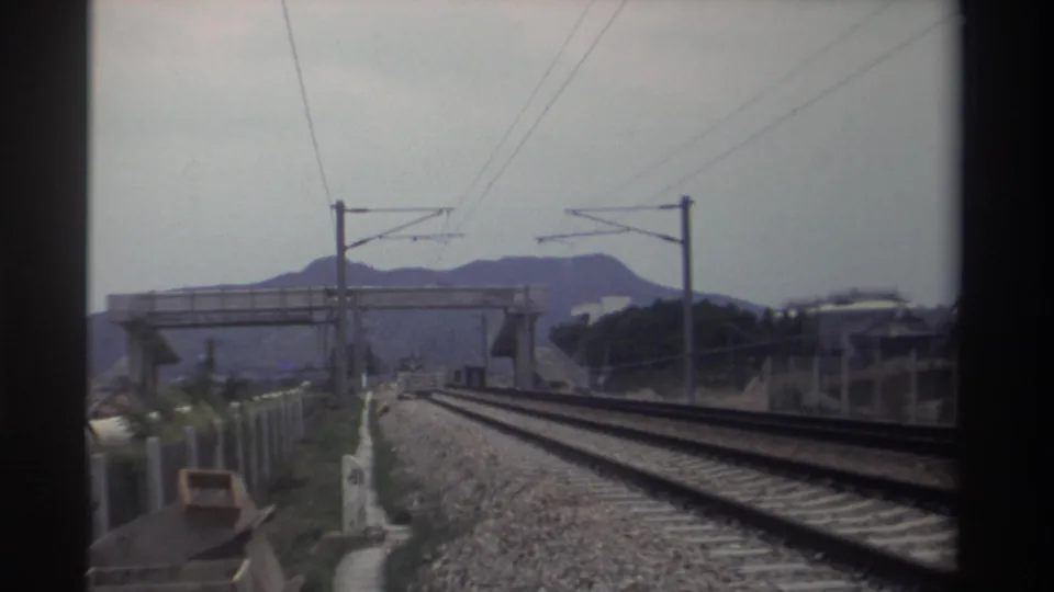 8mm shot looking ahead along dual electrified tracks in Taiwan, 1982, with catenary poles, a concrete overpass, and a mountain ridge visible.