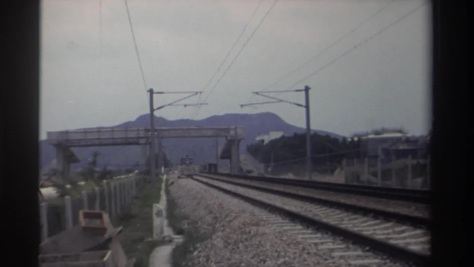 8mm footage of electrified dual railway tracks in Taiwan, 1982; concrete overpass and layered mountain silhouette visible under overcast sky.