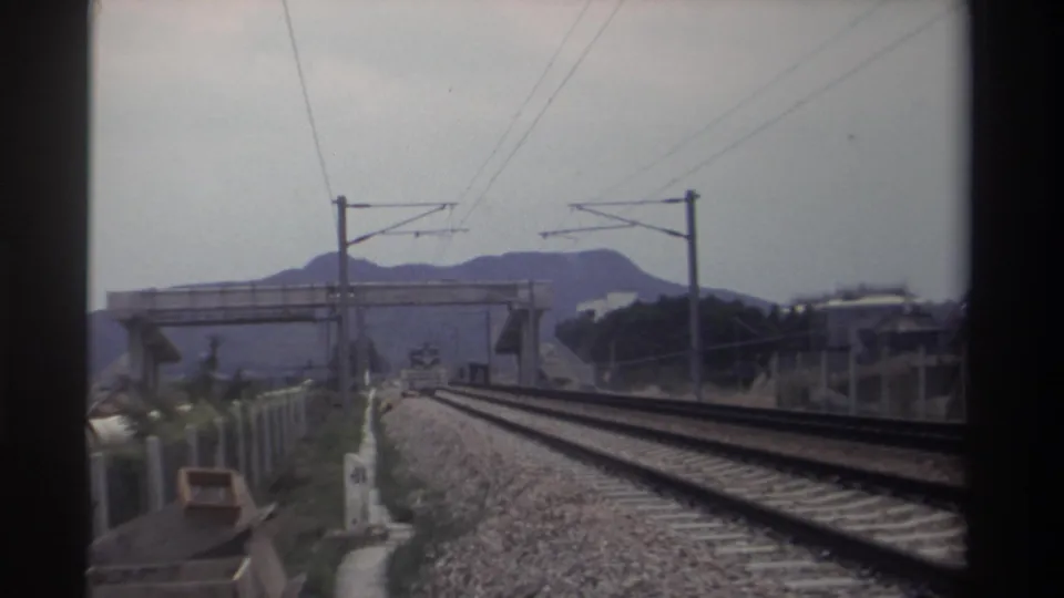 Late-clip 8mm frame of Taiwan railway corridor, 1982; catenary wires span dual tracks leading to a pedestrian overpass with mountain ridge behind.