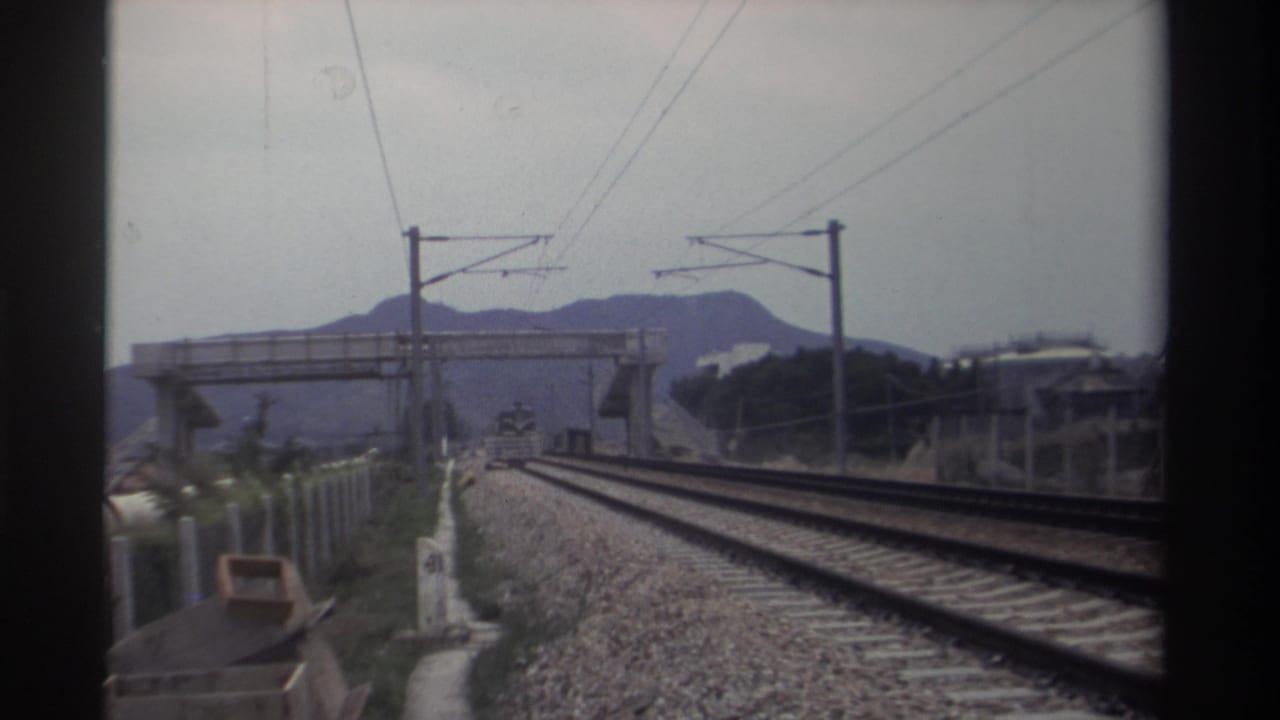 Railway tracks receding ahead from a moving train, with electric catenary gantries and a concrete overpass, mountains in background.