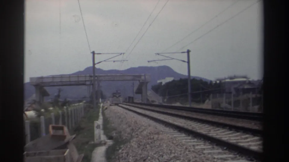 8mm home movie, Taiwan 1982 — forward-looking view from a moving train showing electrified double-track railway, catenary wires, pedestrian overpass, and distant hills.