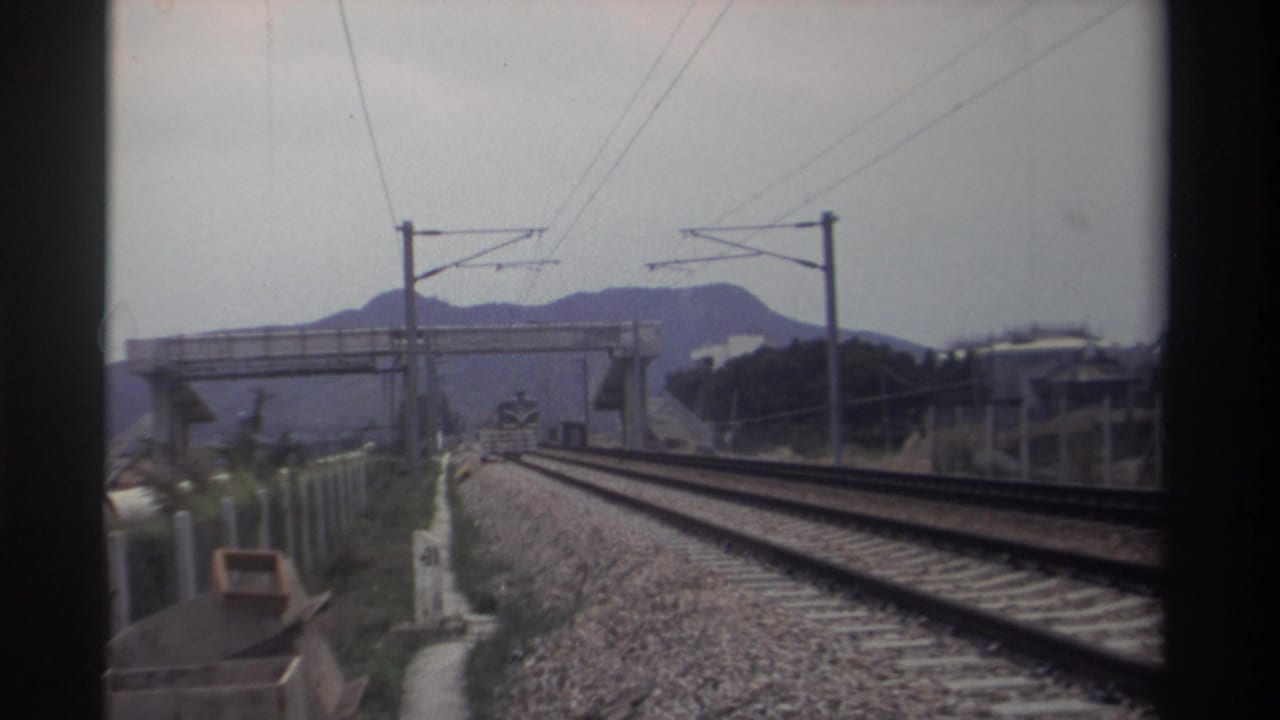 Moving-train POV along double electrified railway tracks with catenary wires, pedestrian overpass, and mountains visible ahead.