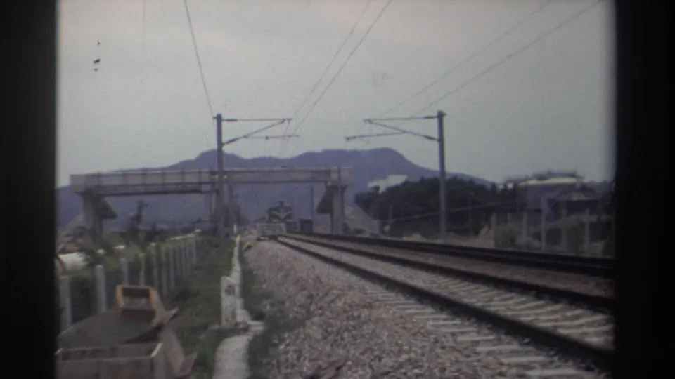8mm home-movie frame, Taiwan 1982 — electrified track corridor with catenary gantries, a concrete footbridge, and hilly ridgeline in diffuse overcast light.
