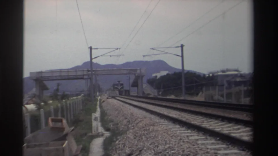 8mm footage, Taiwan 1982 — closing frame of train-window sequence showing parallel tracks, overhead electric lines, pedestrian bridge, and distant hills in flat grey light.