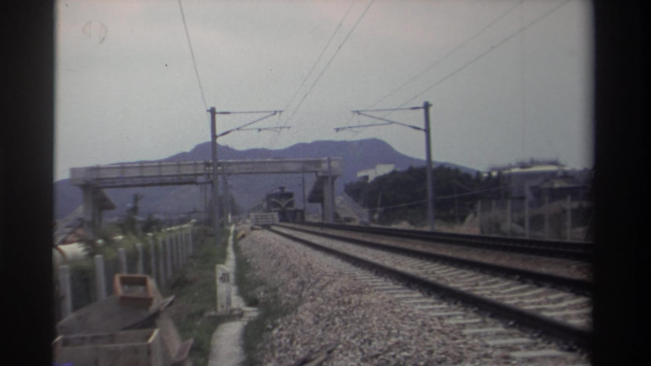 Railway tracks stretching toward a concrete pedestrian overbridge, overhead electrification lines, and hazy mountains under an overcast sky; no train yet visible.