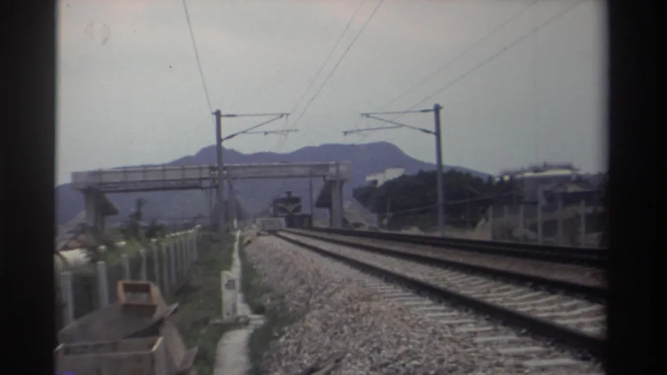 View along an electrified railway in Taiwan, circa 1982, showing double tracks, a concrete pedestrian overbridge, catenary poles, and hazy mountains; 8mm home movie.