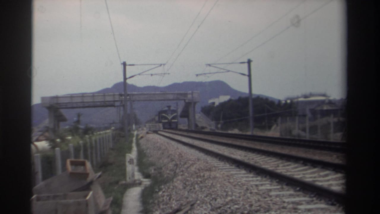 Electrified railway in Taiwan, circa 1982, with a distant blue train approaching below a concrete overbridge, catenary poles, and hazy mountains; 8mm home movie footage.