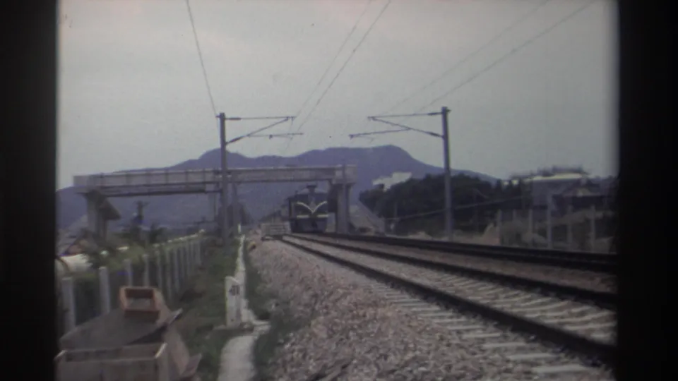 Blue train approaching at mid-distance on an electrified line in Taiwan, circa 1982, with a concrete overbridge, catenary poles, and hazy mountains visible; 8mm footage.