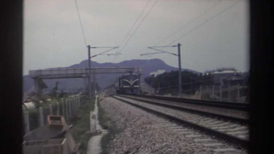 Blue electric train approaching close on an electrified railway in Taiwan, circa 1982; concrete overbridge receding, catenary wires overhead, mountains behind; 8mm home movie.