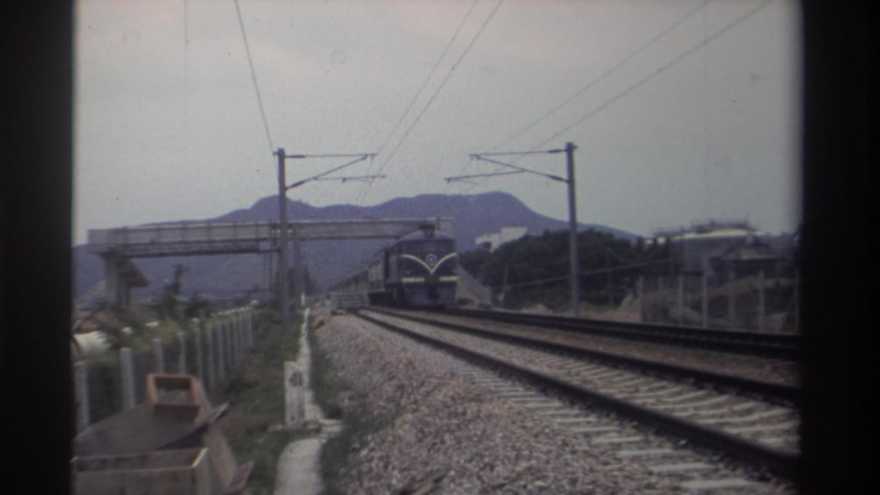 8mm home movie frame showing a locomotive approaching on a double-track electrified line in Taiwan, circa 1982, with a pedestrian overpass and mountain ridgeline behind.