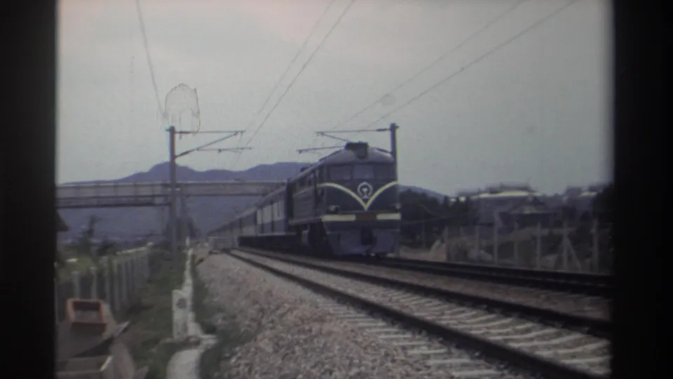 Close-medium 8mm frame of a Taiwan Railways locomotive circa 1982 showing white chevron front markings and an institutional circular emblem; trailing carriages and catenary wires visible.
