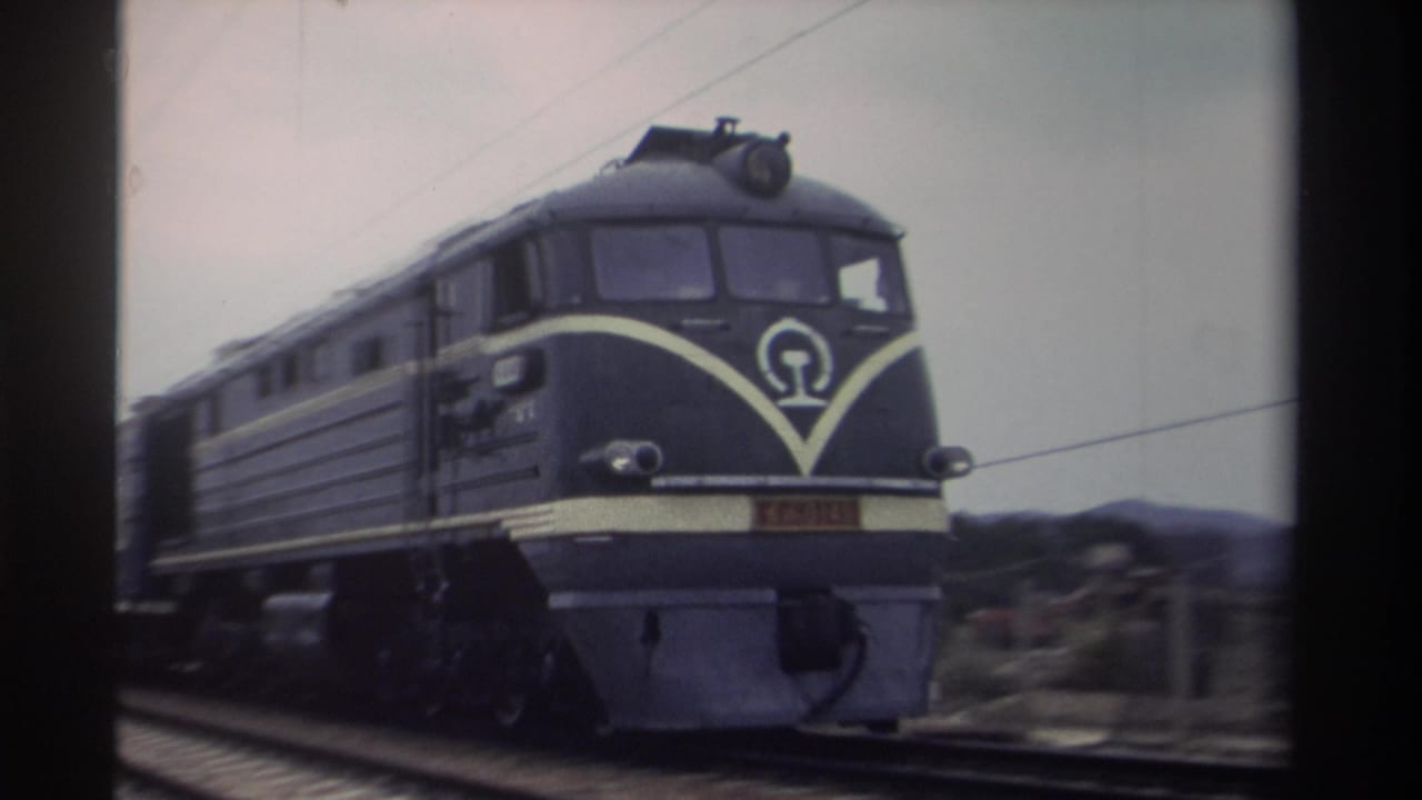 Taiwan Railways-style dark navy locomotive approaching on electrified track, circular authority logo on nose, 8mm home movie grain, Taiwan 1982.