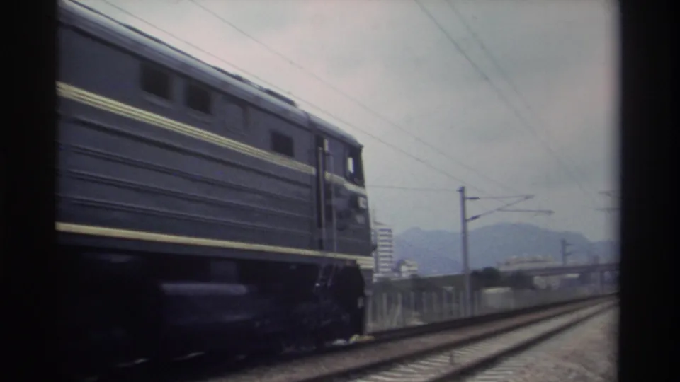 Taiwan Railways dark navy locomotive and carriages passing in profile on electrified line, hazy mountains and suburban structures behind, 8mm film 1982.
