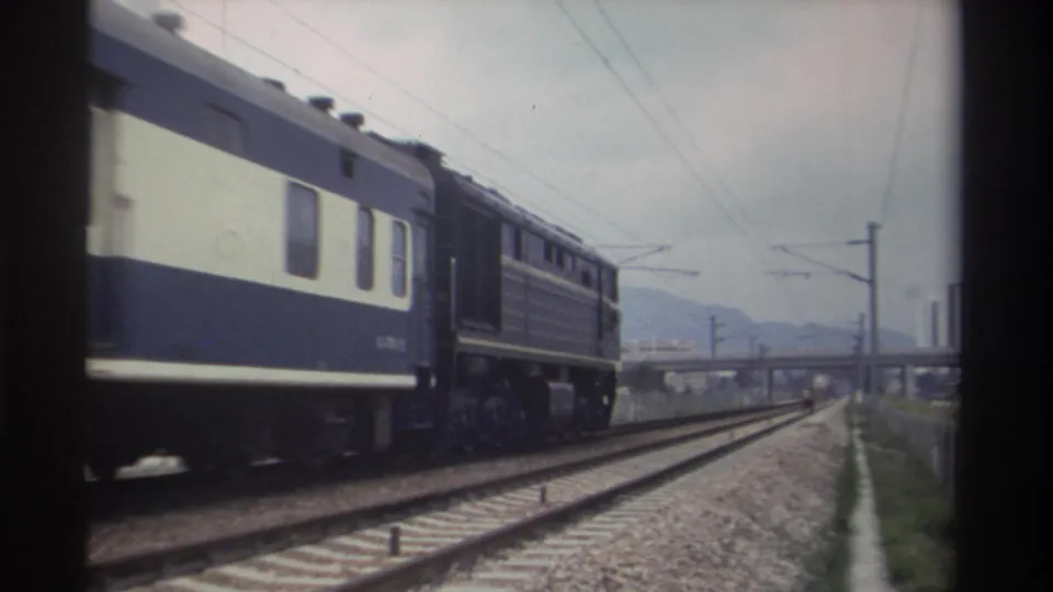 Taiwan Railways carriages receding past catenary poles toward a distant overpass, hazy hills visible; 8mm home movie, Taiwan 1982.