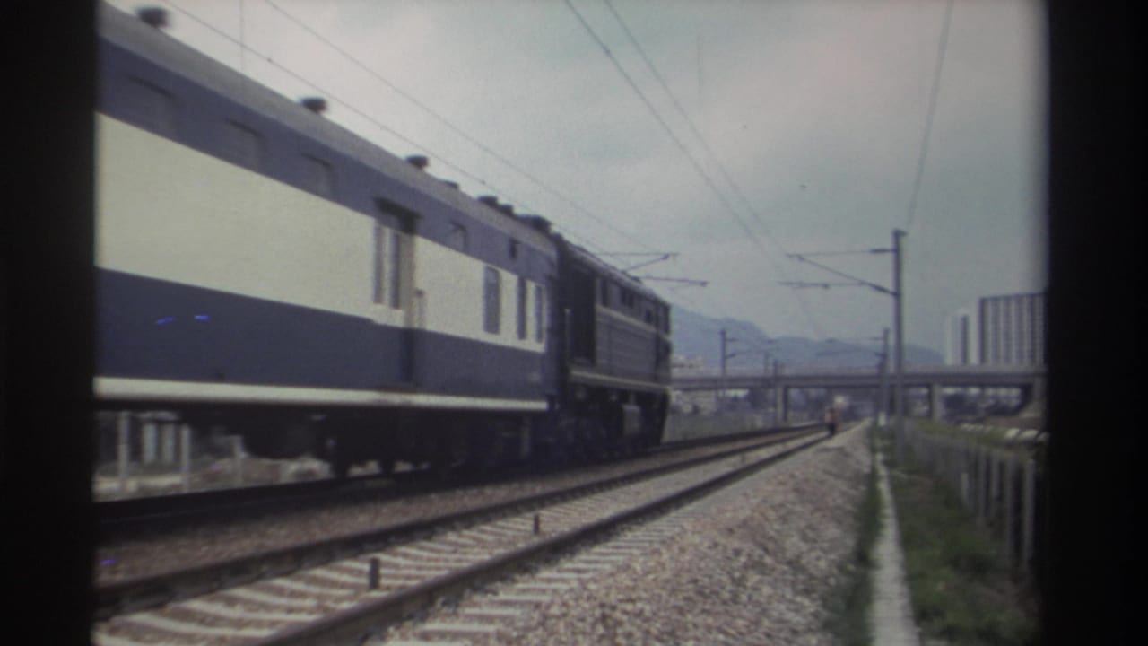 Blue-and-cream passenger train approaching on an electrified track in Taiwan, circa 1982, with a concrete overpass and distant high-rise buildings in the background.
