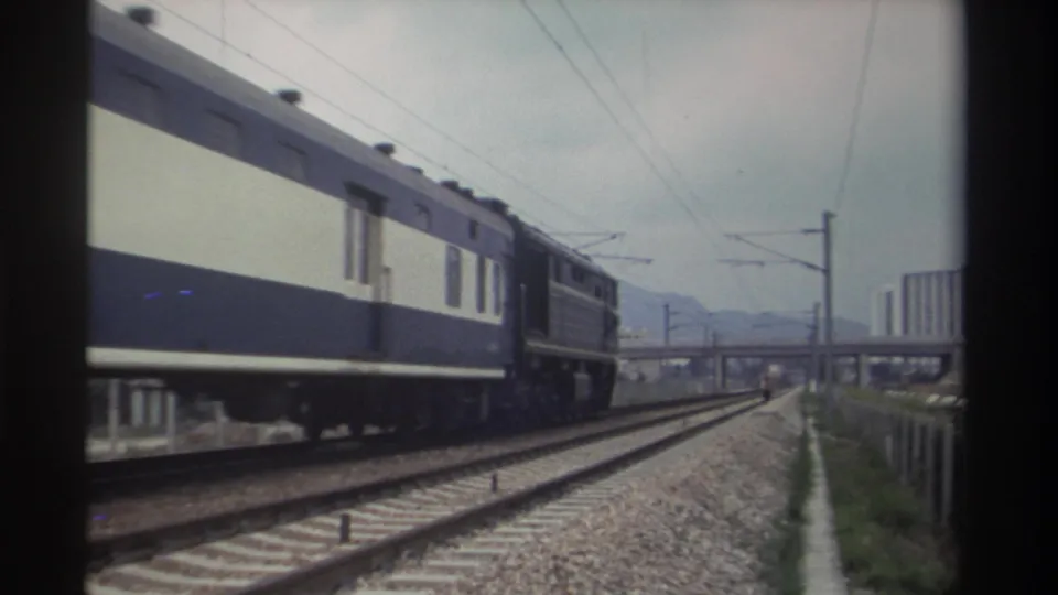 Blue-and-cream passenger train approaching on an electrified track in Taiwan, circa 1982, with a concrete overpass and distant high-rise buildings in the background.