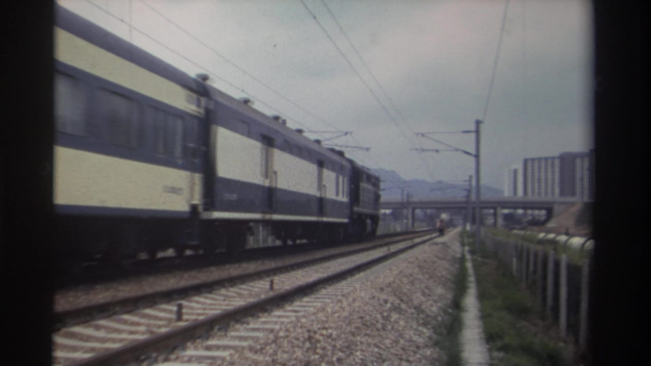 Side view of blue-and-cream passenger train cars on an electrified railway in Taiwan, circa 1982, with residential high-rises and distant mountains visible in the background.