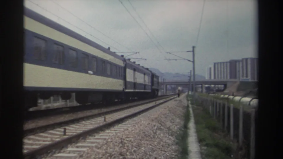 Blue-and-cream passenger train on electrified railway in Taiwan, circa 1982; a small pedestrian figure is visible near the concrete overpass in the mid-ground background.