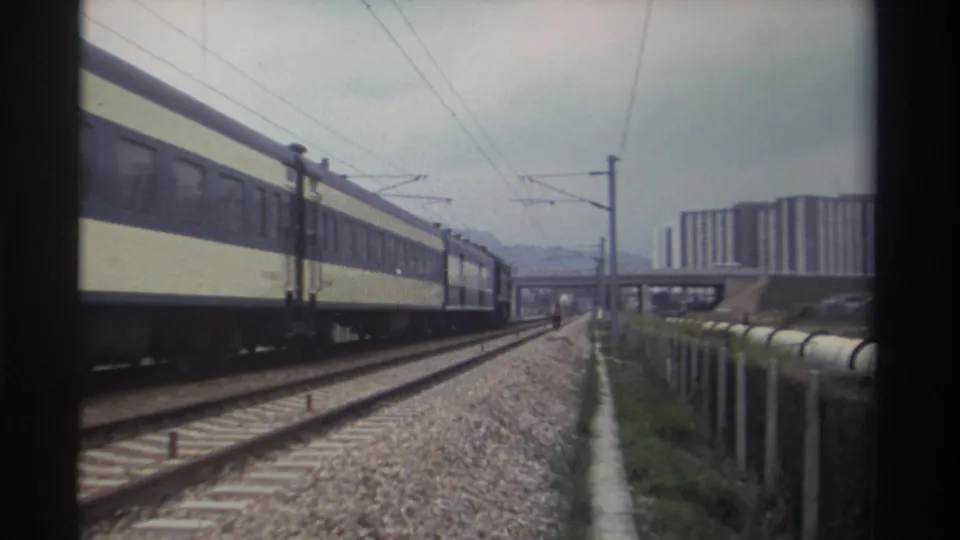 Rear of a blue-and-cream passenger train receding along an electrified railway in Taiwan, circa 1982, with a concrete overpass and residential buildings in the background.