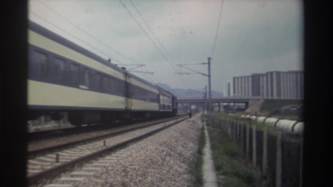 8mm home movie frame, c. 1982, Taiwan — tan-and-dark passenger train on electrified multi-track line, mid-rise apartment blocks and elevated roadway in background.