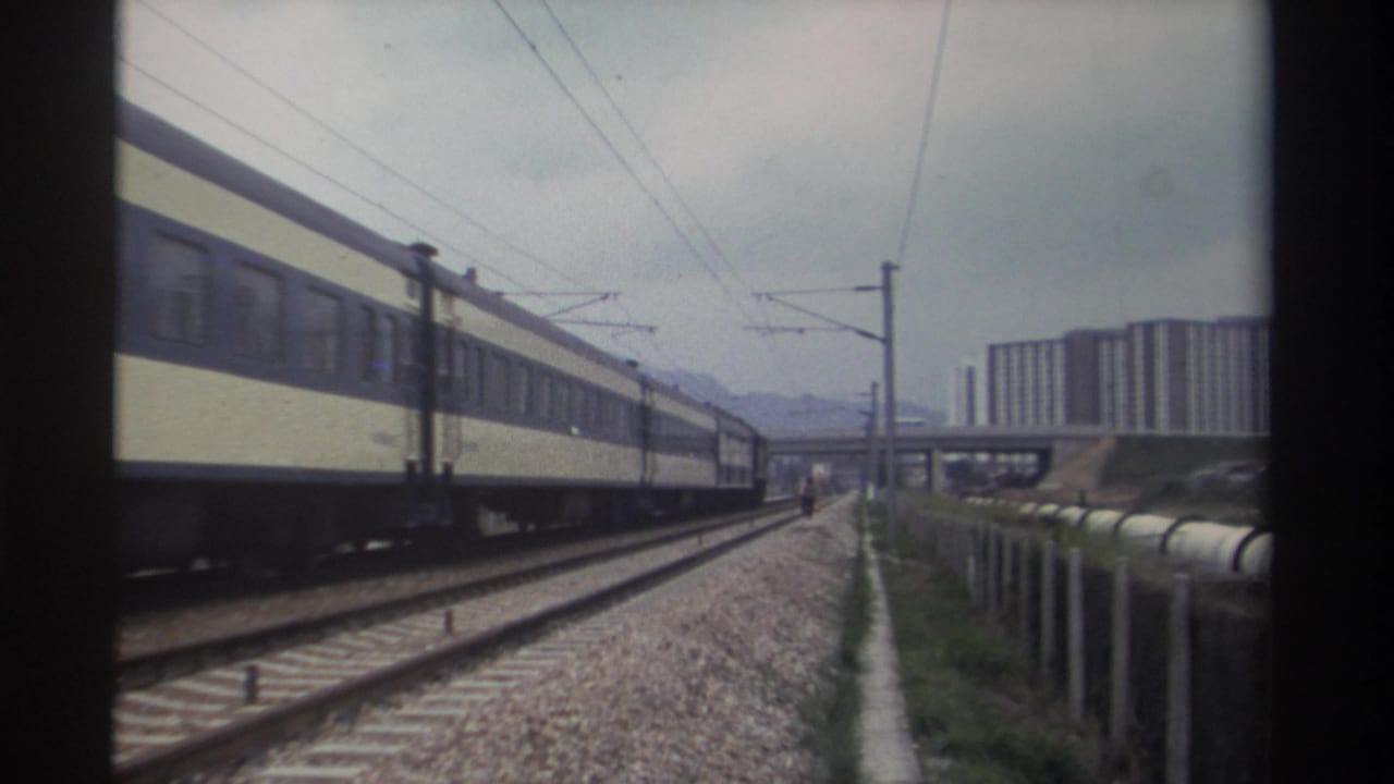 8mm archival frame, c. 1982, Taiwan — electrified passenger train moving alongside multi-track corridor; concrete apartment blocks and overpass visible in mid-distance.