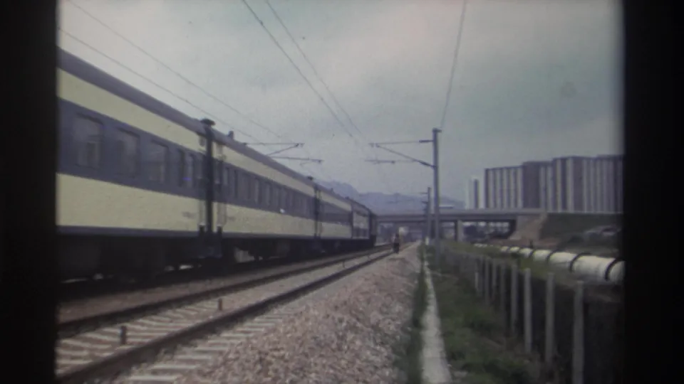 8mm archival frame, c. 1982, Taiwan — electrified passenger train moving alongside multi-track corridor; concrete apartment blocks and overpass visible in mid-distance.
