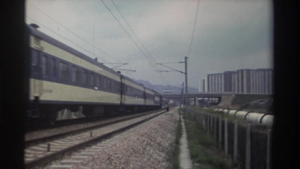 8mm home movie frame, c. 1982, Taiwan — locomotive end of passenger train at left, electrified corridor and concrete housing blocks in background under overcast sky.