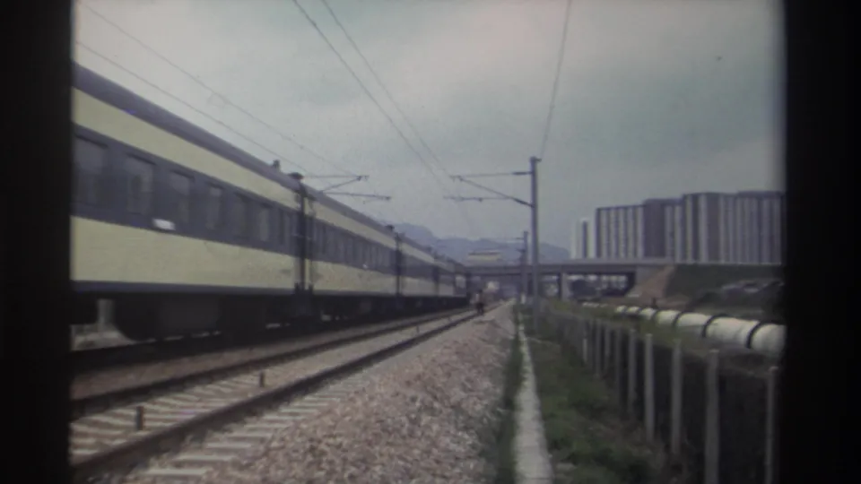8mm archival frame, c. 1982, Taiwan — tan-and-dark passenger train on electrified line, distant apartment towers and overpass visible, characteristic film colour fade.