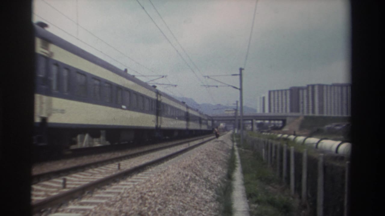 Yellow and blue passenger train on electrified multi-track rail corridor, concrete apartment buildings in background, overcast sky.